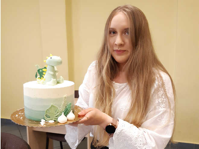 uczennica prezentuje na egzaminie sprawdzającym tort wykończony techniką malowania, udekorowany figurką dinozaura z masy cukrowej
