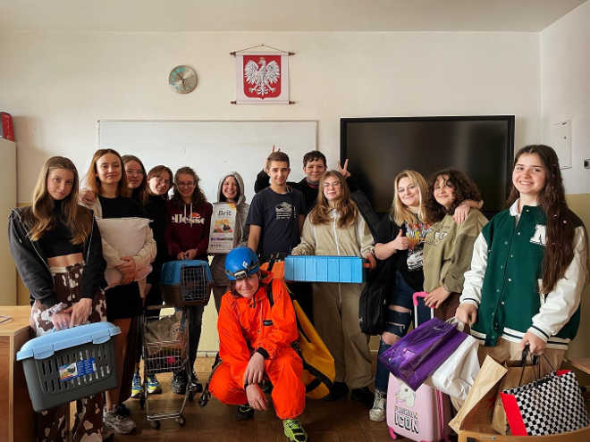 Grupa uśmiechniętych uczniów i nauczycieli, trzymających swoje rzeczy w kubłach, torbach, walizkach i pudełkach, koszach na śmieci, reklamówkach, skrzynkach